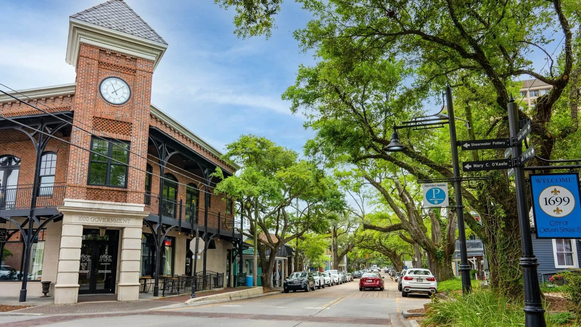 Historic downtown Ocean Springs under the live oak canopy, with the clock tower and the 1699 City of Ocean Springs welcome sign